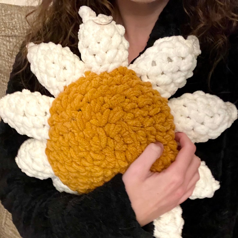 May include: A crocheted sunflower pillow with a white center and yellow petals. The pillow is being held by a person.