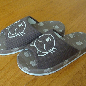 May include: A pair of brown and white slippers with a cat face design. The slippers have a white trim and a paw print pattern.