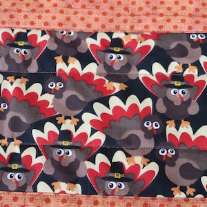 May include: A black fabric with a repeating pattern of brown turkeys wearing pilgrim hats. The turkeys have red, white, and beige feathers. The fabric has a brown border with orange polka dots.