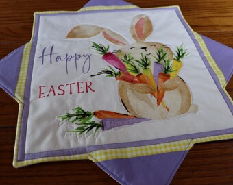 May include: A white fabric Easter table runner with a purple and yellow checkered border. The center features a watercolor illustration of a bunny holding carrots with the text "Happy Easter" in pink and purple.