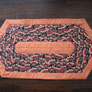 May include: A hexagonal table runner with a Thanksgiving theme. The runner is orange with white polka dots and features a repeating pattern of brown turkeys with red and yellow accents.