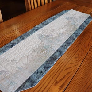 May include: A rectangular table runner with a quilted design. The center is white with a subtle pattern, bordered by a blue and gray patterned fabric. The runner is placed on a wooden table, showcasing its decorative appeal.