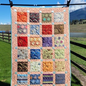 Könnte beinhalten: Eine Patchwork-Decke mit einer Vielzahl von bunten Stoffen und Mustern. Die Decke hängt an einer Wäscheleine im Freien vor dem Hintergrund eines blauen Himmels und grünen Grases.