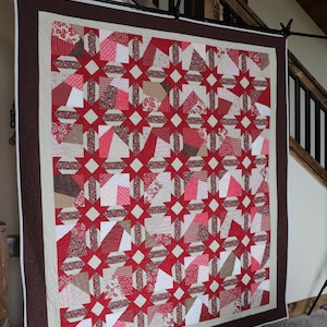 以下が含まれることがあります： 赤、白、タンの色合いの星柄の手作りキルト。キルトは茶色の縁取りがあり、黒い棒に展示されています。デザインにはさまざまな生地パターンが含まれており、居心地の良い装飾的なテキスタイルを作成しています。