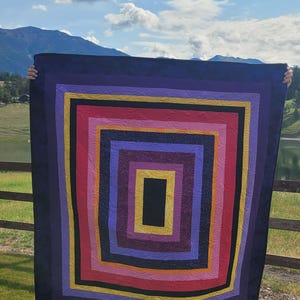 May include: A colorful, geometric quilt with a nested rectangle design. The quilt features a dark blue border, surrounding layers of purple, yellow, red, and orange. The center is a black rectangle. The quilt is held up against a backdrop of mountains and a lake.