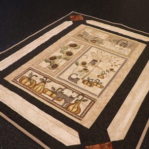 Puede incluir: Un edredón rectangular con temática otoñal. El edredón presenta paneles con girasoles, calabazas y peras. El borde es negro con detalles en beige y marrón. El edredón está sobre una superficie oscura.