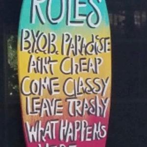 May include: A surfboard-shaped sign with the words "POOL RULES" painted in white on a turquoise background. The sign also has the text "BYOB PARADISE AINT CHEAP COME CLASSY LEAVE TRASHY WHAT HAPPENS HERE STAYS HERE" painted in white on a yellow, orange, and red background.