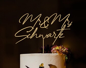Puede incluir: Topper de pastel dorado con el texto "Mr & Mrs  Schnark" en una fuente cursiva.