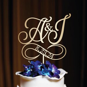 May include: Gold wedding cake topper with the letters A & J in a cursive font and the date 8.23.24 on a ribbon.