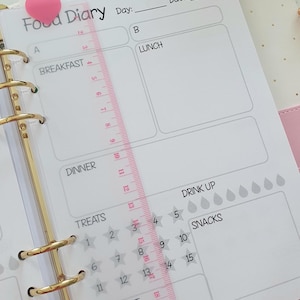 May include: A white food diary planner with a pink heart sticker on the cover. The planner has sections for breakfast, lunch, dinner, treats, snacks, drink up, and workout. There is a ruler on the side of the planner.