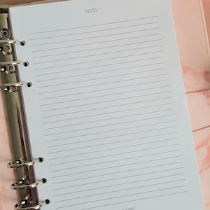 Könnte beinhalten: Ein weißes, liniertes Papier-Notizbuch mit einem silbernen Binder und dem Wort "NOTES" oben aufgedruckt.