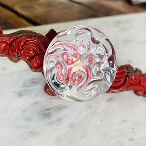 May include: A clear glass knob with a textured surface, set on a red, ornate, metal base. The base has a detailed, scroll-like design. The knob is round and reflects light, creating a swirling pattern.