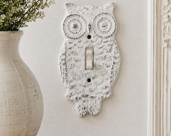 May include: A white owl-shaped light switch cover with a distressed, weathered finish. The cover features detailed feather textures and two circular eyes, with a central rectangular cutout for a standard toggle switch and two screw holes.