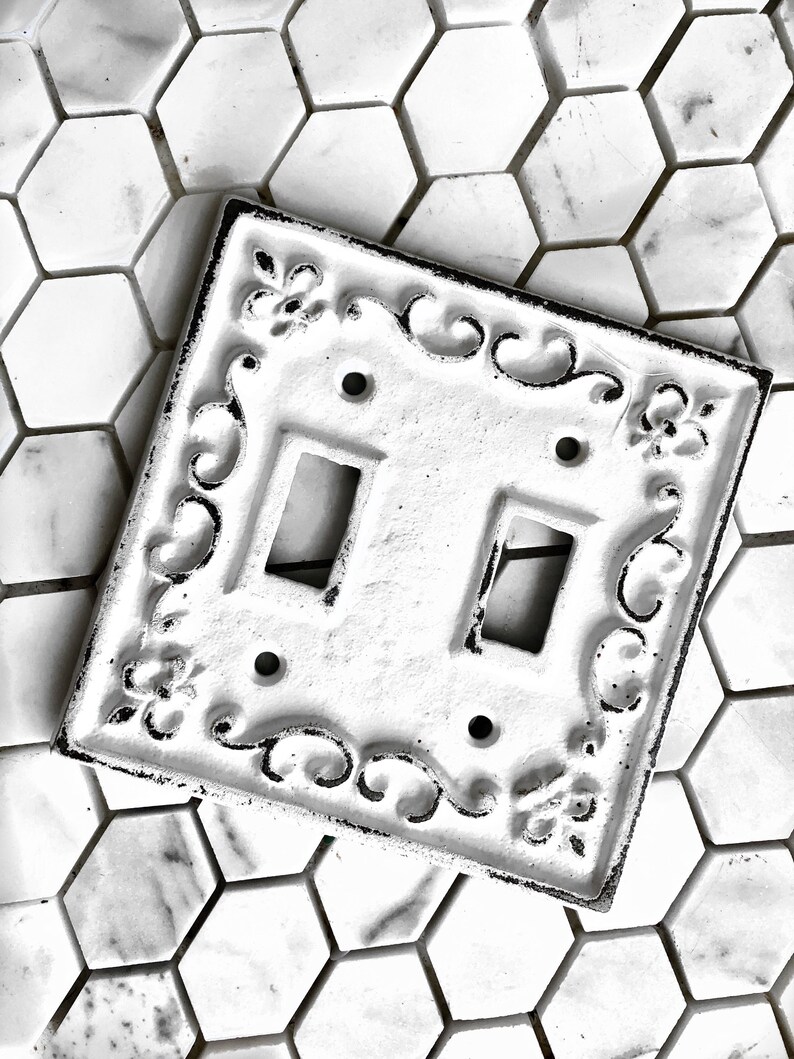 May include: White porcelain double light switch cover with an ornate black design. The switch cover is on a white hexagonal tile background.