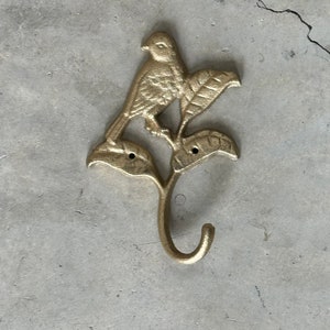 May include: A gold-colored metal wall hook featuring a bird perched on a leafy branch. The hook is curved and positioned below the bird and leaves. The bird and leaves are detailed with textured surfaces.