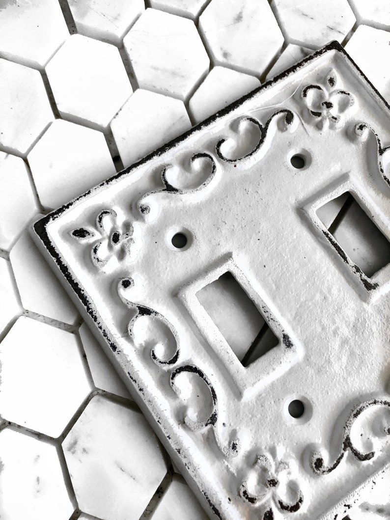 May include: White porcelain light switch cover with an ornate, decorative design. The switch cover has two toggle switches and is accented with black detailing.