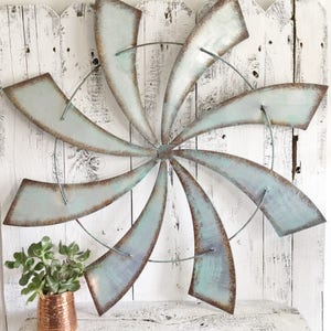 May include: A large metal wall decor in the shape of a pinwheel with eight blades. The blades are painted a light blue-green color with a distressed finish. The pinwheel is mounted on a white wooden wall.