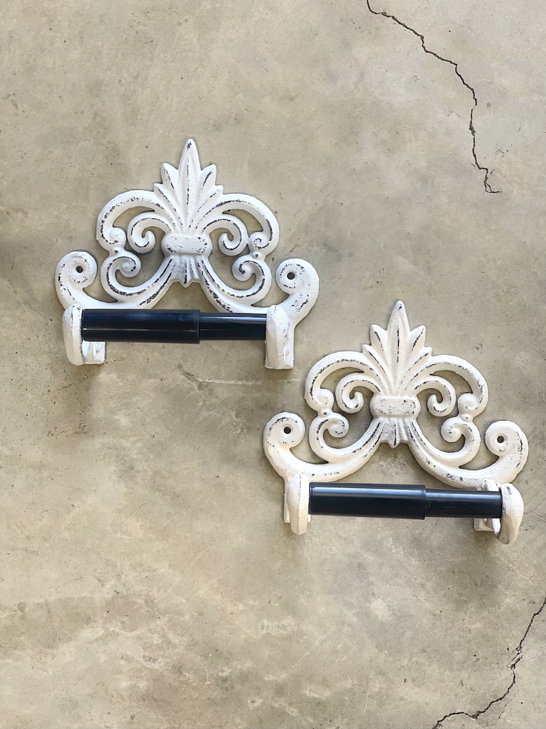 May include: Two white cast iron toilet paper holders with ornate scrollwork designs. The holders are mounted on a textured wall and feature black metal toilet paper rolls.
