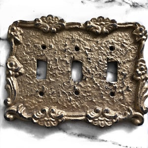 May include: A gold-colored metal light switch plate with an ornate design and three toggle switches.