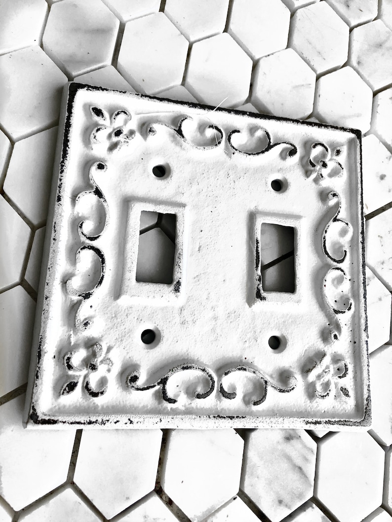 May include: White cast iron double light switch cover with an ornate design. The cover has a distressed finish and is perfect for adding a touch of vintage charm to any room.