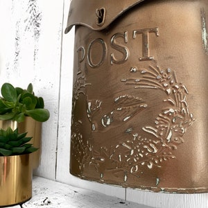 Puede incluir: Un buz&oacute;n de metal de estilo vintage con un acabado dorado desgastado. El buz&oacute;n tiene una solapa con un pestillo de metal y la palabra "POST" en relieve en la parte delantera. El buz&oacute;n est&aacute; decorado con un dise&ntilde;o floral.