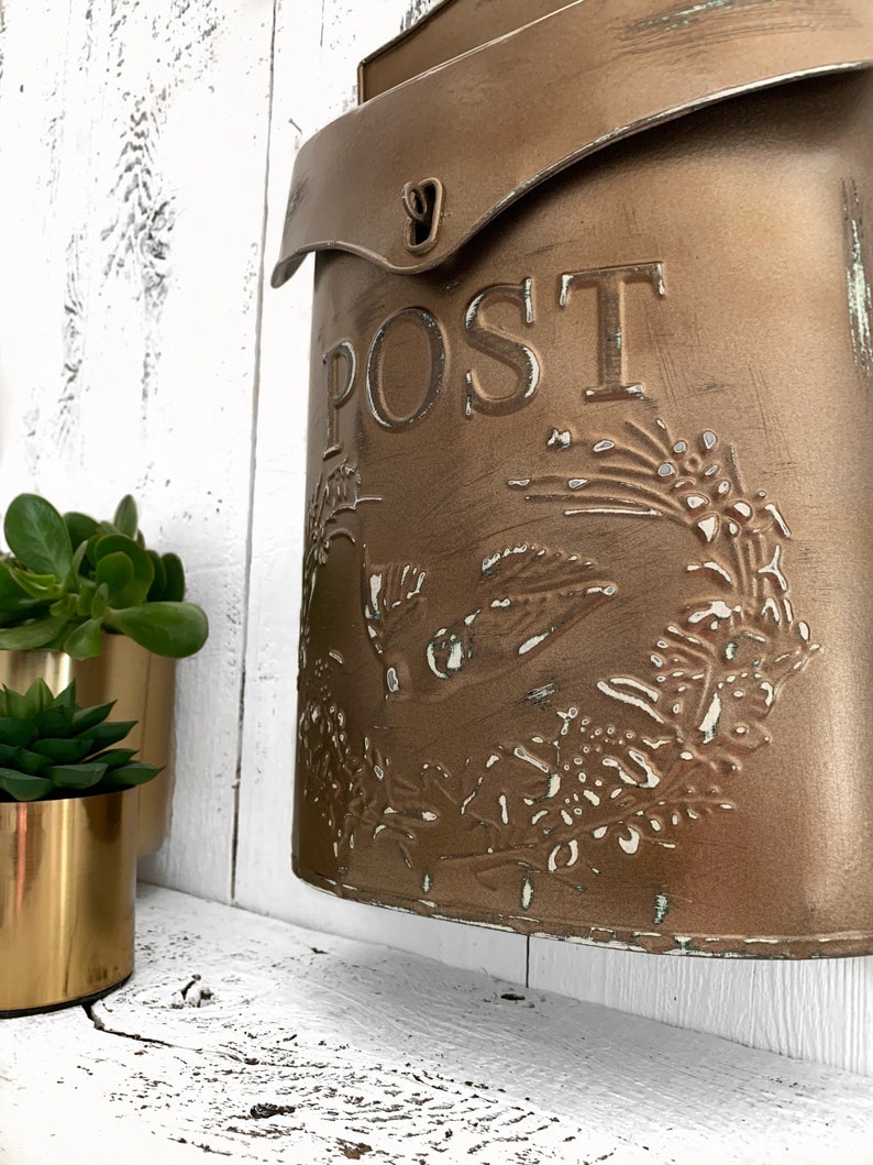 Puede incluir: Un buz&oacute;n de metal de estilo vintage con un acabado dorado desgastado. El buz&oacute;n tiene una solapa con un pestillo de metal y la palabra "POST" en relieve en la parte delantera. El buz&oacute;n est&aacute; decorado con un dise&ntilde;o floral.