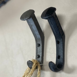 May include: Two black metal hooks shaped like nails with rounded heads. The hooks are mounted on a white wall and a piece of twine is hanging from one of the hooks.