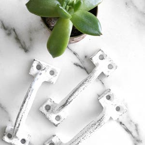 May include: Three white cast iron drawer pulls with a distressed finish. The pulls have a square backplate and a curved handle.