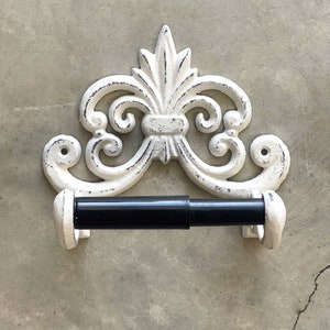 May include: White cast iron toilet paper holder with a decorative scroll design. The holder has a black metal rod for holding the toilet paper.