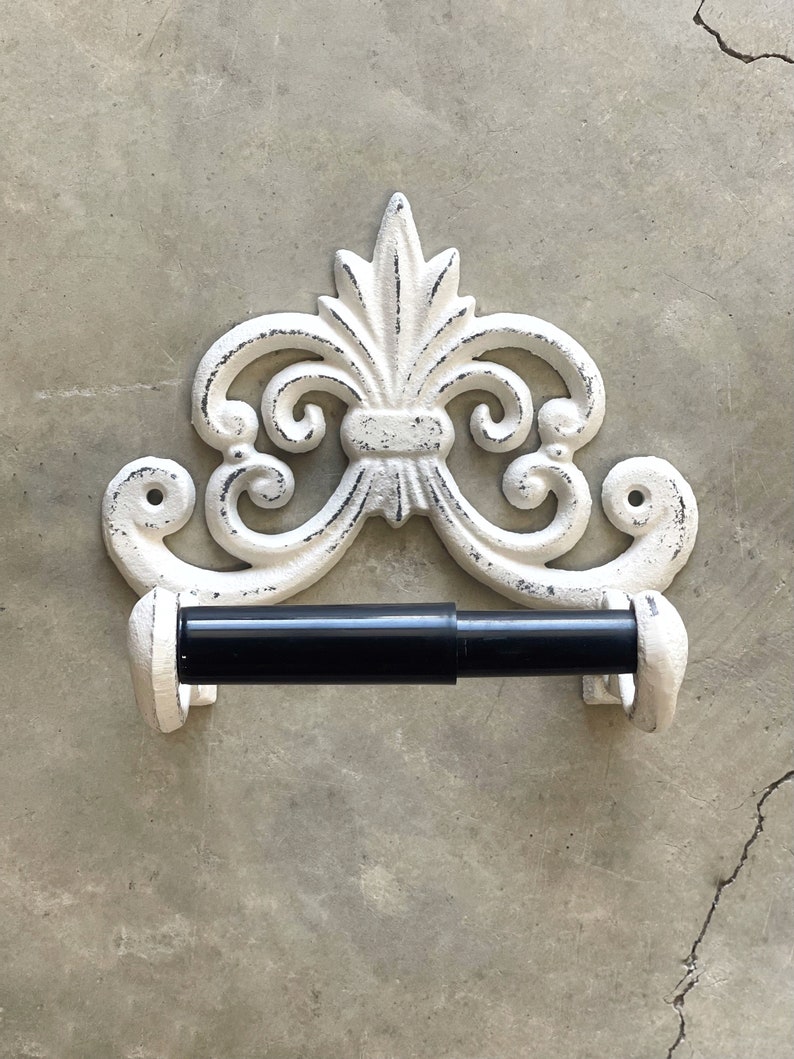 May include: White cast iron toilet paper holder with a decorative scroll design. The holder has a black metal rod for holding the toilet paper.