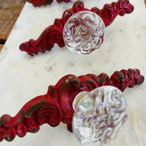 Puede incluir: Juego de tres tiradores de armario de estilo antiguo. Cada tirador presenta un pomo de cristal transparente texturizado y una base de metal roja ornamentada. La base met&aacute;lica tiene un acabado envejecido, lo que a&ntilde;ade a la est&eacute;tica vintage.