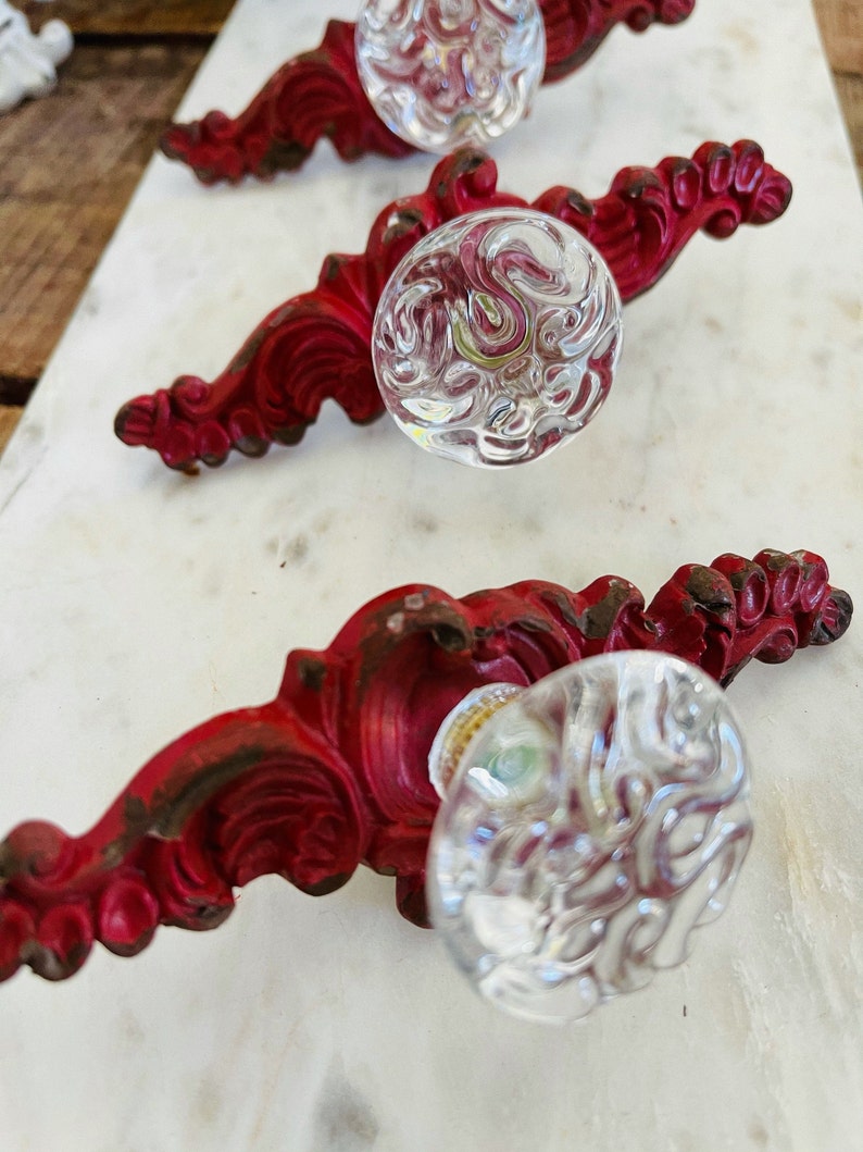 Puede incluir: Juego de tres tiradores de armario de estilo antiguo. Cada tirador presenta un pomo de cristal transparente texturizado y una base de metal roja ornamentada. La base met&aacute;lica tiene un acabado envejecido, lo que a&ntilde;ade a la est&eacute;tica vintage.