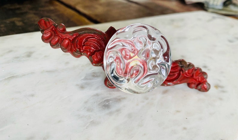 Puede incluir: Un pomo de cristal transparente con una superficie texturizada, colocado sobre una base met&aacute;lica roja y ornamentada. La base tiene un dise&ntilde;o detallado en forma de voluta. El pomo es redondo y refleja la luz.