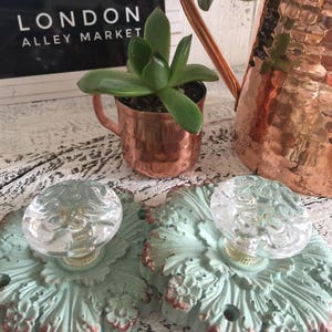 May include: Two decorative door knobs with a distressed, light green finish and ornate details. Each knob features a clear glass knob with a floral design. The knobs are displayed on a white wooden surface.
