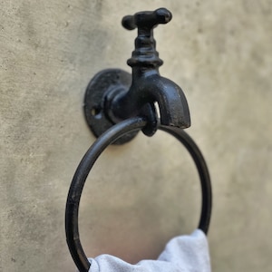 May include: Black cast iron towel ring with a faucet design. The ring is mounted on a wall and has a towel hanging from it.