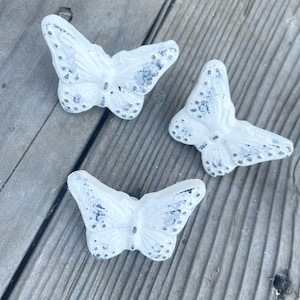 May include: Three white ceramic butterfly figurines with black accents. The butterflies are arranged on a wooden surface.