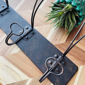 May include: A black metal coat rack with three hooks. The rack has a rustic, distressed finish. The rack is mounted on a wooden surface with a chevron pattern.