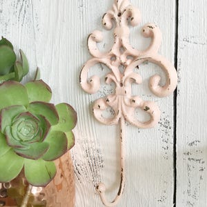 May include: A pink metal hook with a decorative, ornate design. The hook is mounted on a white wooden wall.