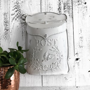 May include: White metal mailbox with a floral design and the word "POST" on the front. The mailbox is mounted on a white wooden wall.