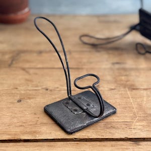 May include: A black metal hook with a square base. The hook is attached to the base with two metal rivets. The hook is bent into a curved shape, with a small loop at the top.