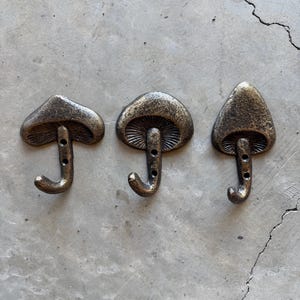 May include: Three bronze-colored mushroom-shaped wall hooks. Each hook has a curved bottom for hanging items and a detailed cap design. The hooks are mounted on a gray concrete surface.