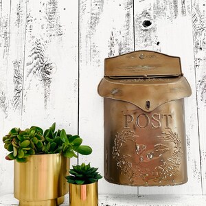 Puede incluir: Un buz&oacute;n vintage dorado con la palabra "POST" en relieve en la parte delantera. El buz&oacute;n est&aacute; colgado en una pared de madera blanca. Dos macetas doradas con suculentas verdes est&aacute;n en primer plano.