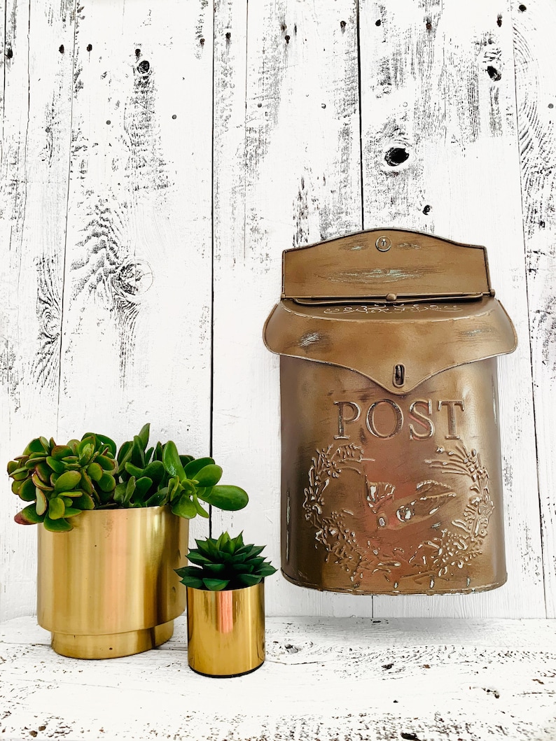 Puede incluir: Un buz&oacute;n vintage dorado con la palabra "POST" en relieve en la parte delantera. El buz&oacute;n est&aacute; colgado en una pared de madera blanca. Dos macetas doradas con suculentas verdes est&aacute;n en primer plano.