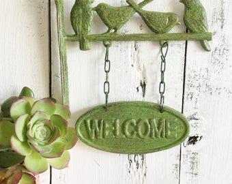 May include: Green metal welcome sign with three birds perched on a branch. The sign hangs from chains and has the word "WELCOME" embossed on it.