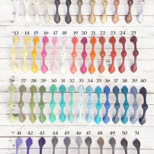 Puede incluir: Una colecci&oacute;n de 51 tiradores decorativos para armarios en varios colores, dispuestos sobre una superficie de madera blanca. Los tiradores tienen un dise&ntilde;o ornamentado y sim&eacute;trico con un elemento decorativo central y numerados del 1 al 51.