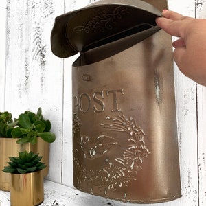Puede incluir: Un buz&oacute;n de metal marr&oacute;n con un dise&ntilde;o floral decorativo y la palabra "POST" en la parte delantera. El buz&oacute;n tiene una tapa con bisagras que est&aacute; abierta.