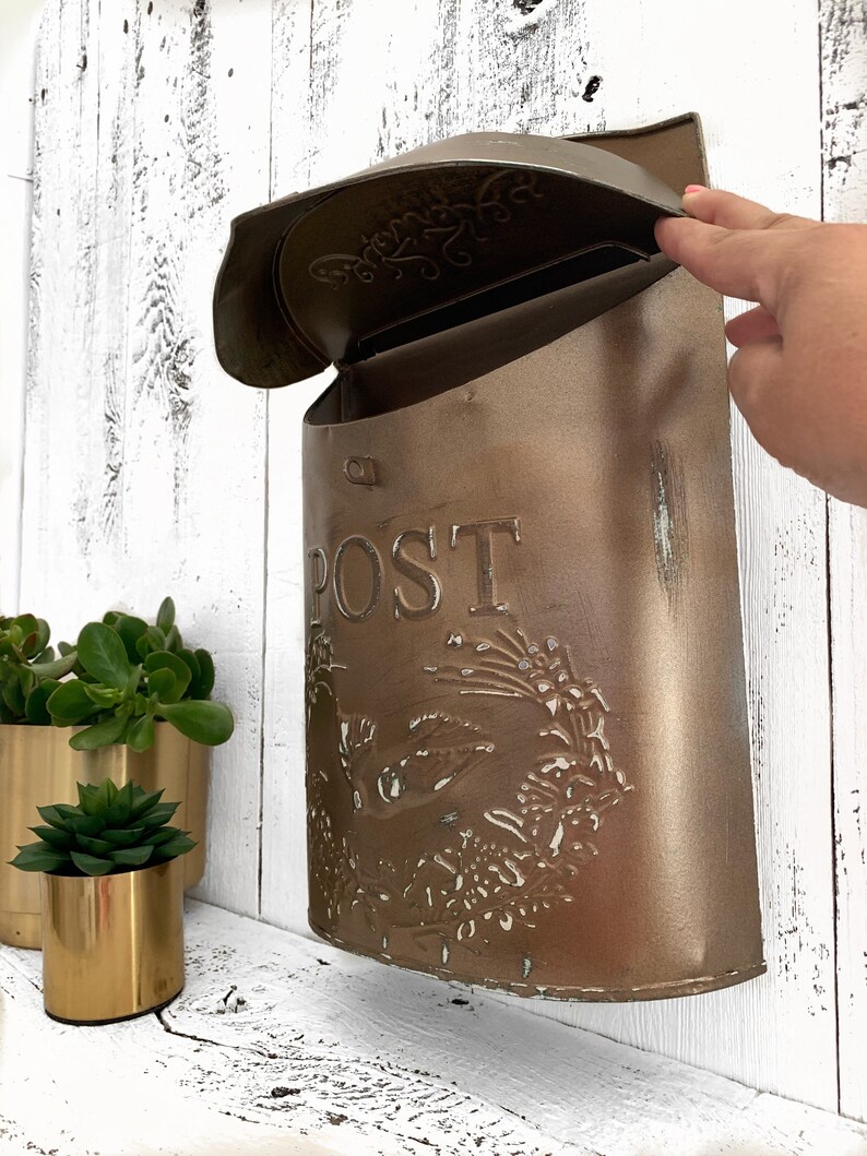 Puede incluir: Un buz&oacute;n de metal marr&oacute;n con un dise&ntilde;o floral decorativo y la palabra "POST" en la parte delantera. El buz&oacute;n tiene una tapa con bisagras que est&aacute; abierta.