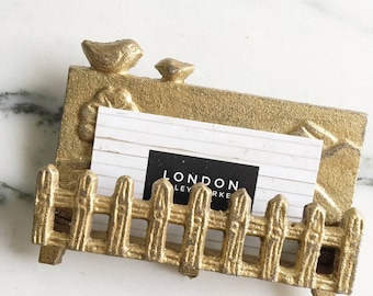May include: Gold metal business card holder shaped like a picket fence with two small birds perched on top. The card holder is sitting on a white marble surface.