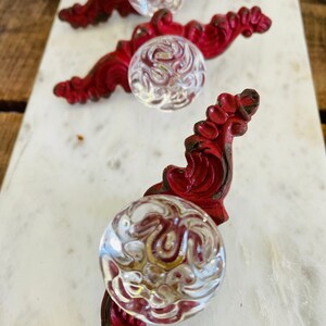 Puede incluir: Tres tiradores de armario de estilo antiguo. Cada tirador presenta un pomo de cristal transparente con dibujos, fijado a una base decorativa de metal pintado de rojo. Las bases tienen un dise&ntilde;o ornamental y enrollado, sobre una superficie de m&aacute;rmol blanco.