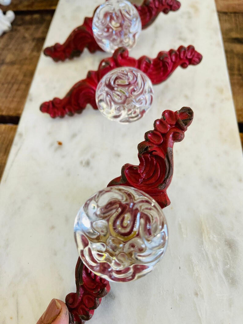 Puede incluir: Tres tiradores de armario de estilo antiguo. Cada tirador presenta un pomo de cristal transparente con dibujos, fijado a una base decorativa de metal pintado de rojo. Las bases tienen un dise&ntilde;o ornamental y enrollado, sobre una superficie de m&aacute;rmol blanco.